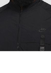 Černá lehká bunda na zip, s vysokým límcem, logem Nike, náprsní kapsou a detailem s nastavitelným páskem. Hladká, matná textura.