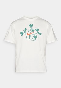 Camiseta blanca de manga corta con hojas de hiedra verdes pixeladas y un logotipo de swish de Nike naranja en el centro frontal.