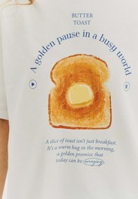 Bílý tričko s grafikou zlatohnědého krajíce toastu s kouskem másla, obklopené modrým textem a hravou typografií.