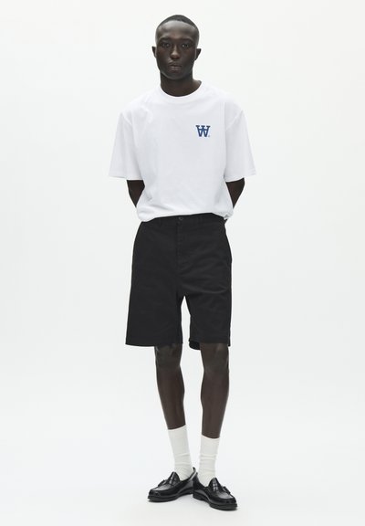 T-shirt blanc à manches courtes avec logo bleu marine, associé à un short noir longueur genoux, des chaussettes blanches et des mocassins noirs. Design et couleurs simples.