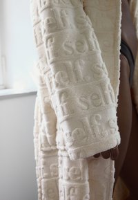 Peignoir couleur crème avec une surface texturée où le mot "self" est embossé en motif répétitif. Matière douce et moelleuse avec manches longues.