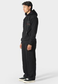 Modelo masculino vistiendo una sudadera negra con bolsillo frontal y pantalones anchos negros a juego, de pie en perfil contra un fondo claro y liso.