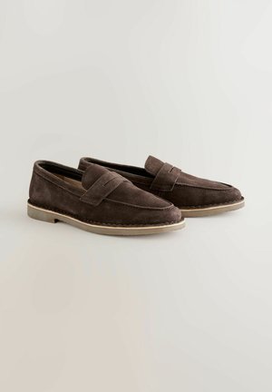 Hnedé semišové mokasíny so šitým vzorom, plochou gumovou podrážkou a výrazným remienkom cez vrch. Hladká textúra a ležérny tvar.