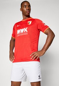 Sarkans futbola krekls ar īsām piedurknēm, ar teksturētu dizainu, balto logotipu un dzeltenām akcentiem. Apvienots ar baltām šortiem un logotipa detaļu.
