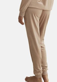 Pantalon de détente beige en matériau doux et velouté. Poignets ajustés aux chevilles. Texture lisse avec une coupe décontractée et sans ornements visibles.