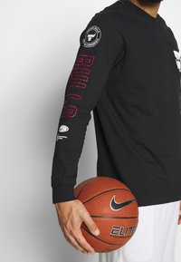 Czarna koszulka z długim rękawem z napisem "BULLS" na rękawie w kolorze czerwonym, z logo Chicago Bulls na klatce piersiowej, zestawiona z piłką do koszykówki.
