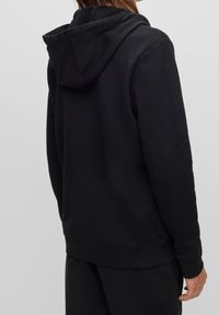 Černá mikina s kapucí a šňůrkou, ribbenými manžetami a lemem, vyrobená z měkké tkaniny s hladkým povrchem. Minimalistický design.