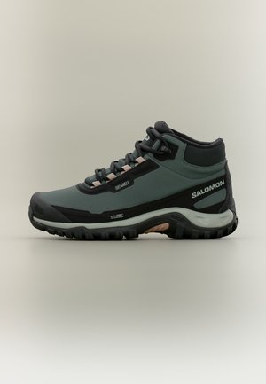 Botte de randonnée mi-hauteur vert foncé et noir avec lacets, semelle texturée et marquage Salomon sur le côté, conçue pour une utilisation extérieure étanche.