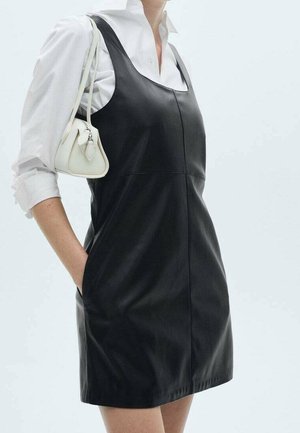 Woman wearing black sleeveless leather dress over white shirt, carrying small white handbag on shoulder, hand in pocket.