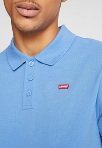 Polo bleu clair en tissu texturé, avec trois boutons et un petit logo rouge Levi's sur la poitrine. Les manches courtes sont visibles.