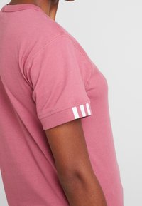 Růžové tričko s krátkým rukávem a kulat ým výstřihem, zdobené bílými pruhy na rukávech. Vyrobeno z měkké bavlny s viditelnými švy.