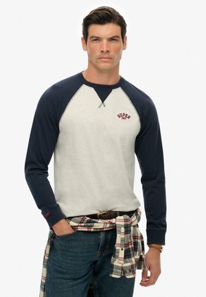 Mężczyzna ubrany w granatowo-szary długi rękaw z logo "Superdry", koszula w kratę zawiązana wokół talii oraz niebieskie dżinsy na białym tle.