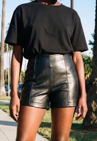Personne portant une chemise noire à manches courtes et un short en cuir noir taille haute, debout à l'extérieur près des palmiers.