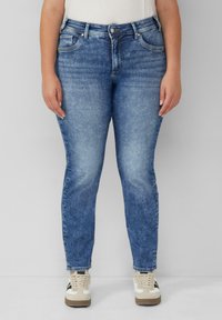 Osoba w niebieskich, wyblakłych dżinsach prostych i białych sneakersach z czarnymi i beżowymi akcentami, stojąca na tle bez wzorów.