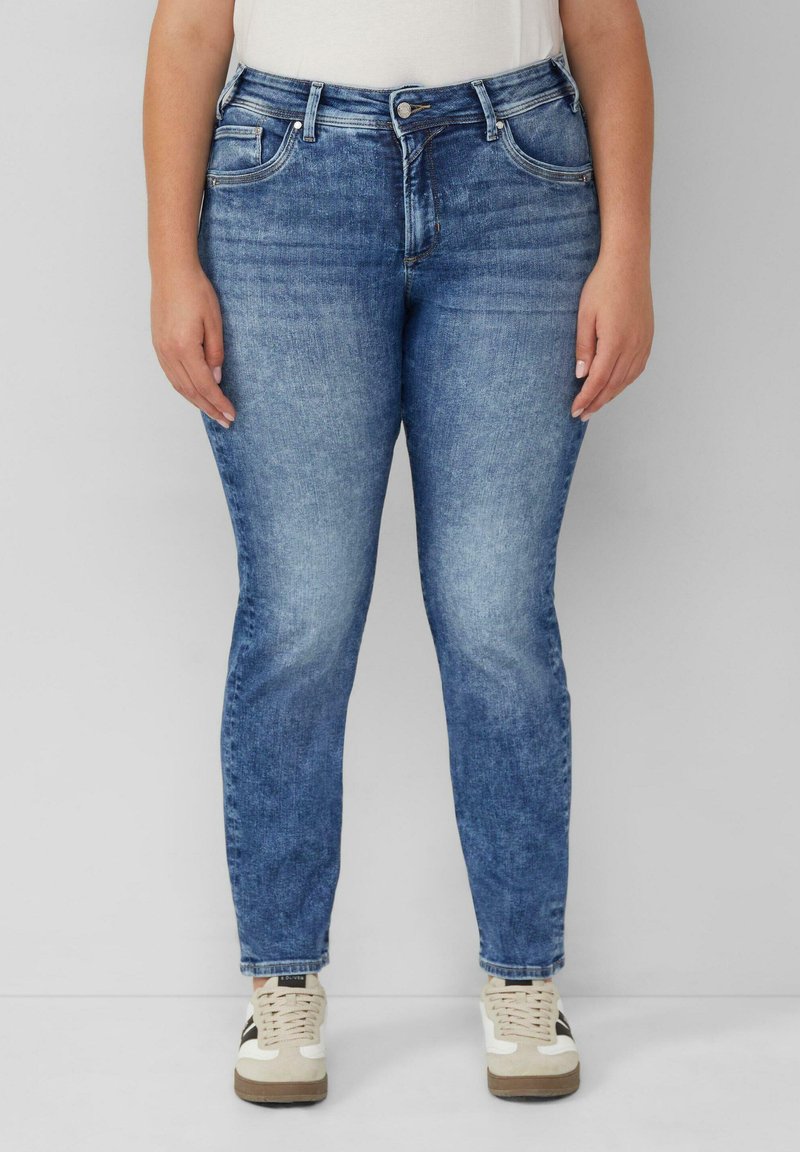 Osoba w niebieskich, wyblakłych dżinsach prostych i białych sneakersach z czarnymi i beżowymi akcentami, stojąca na tle bez wzorów.