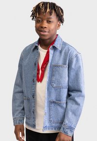 Denim jacket in light blue, featuring a button closure, two front pockets, and contrasting stitching. Underneath, a white shirt with a red bandana.