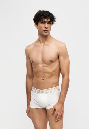 Jeune homme musclé aux cheveux bouclés et foncés, portant un boxer blanc avec une ceinture dorée à motifs, debout sur un fond uni.
