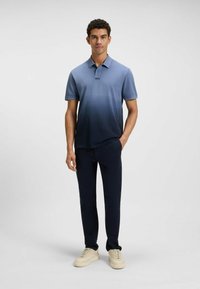 Man står med handen i fickan iförd en blå gradientpolo, mörka byxor och beige sneakers mot en enfärgad ljus bakgrund.