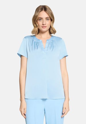 Frau mit schulterlangem blondem Haar, die eine hellblaue kurzärmelige Bluse und passende hellblaue Hose trägt, vor einem schlichten Hintergrund stehend.