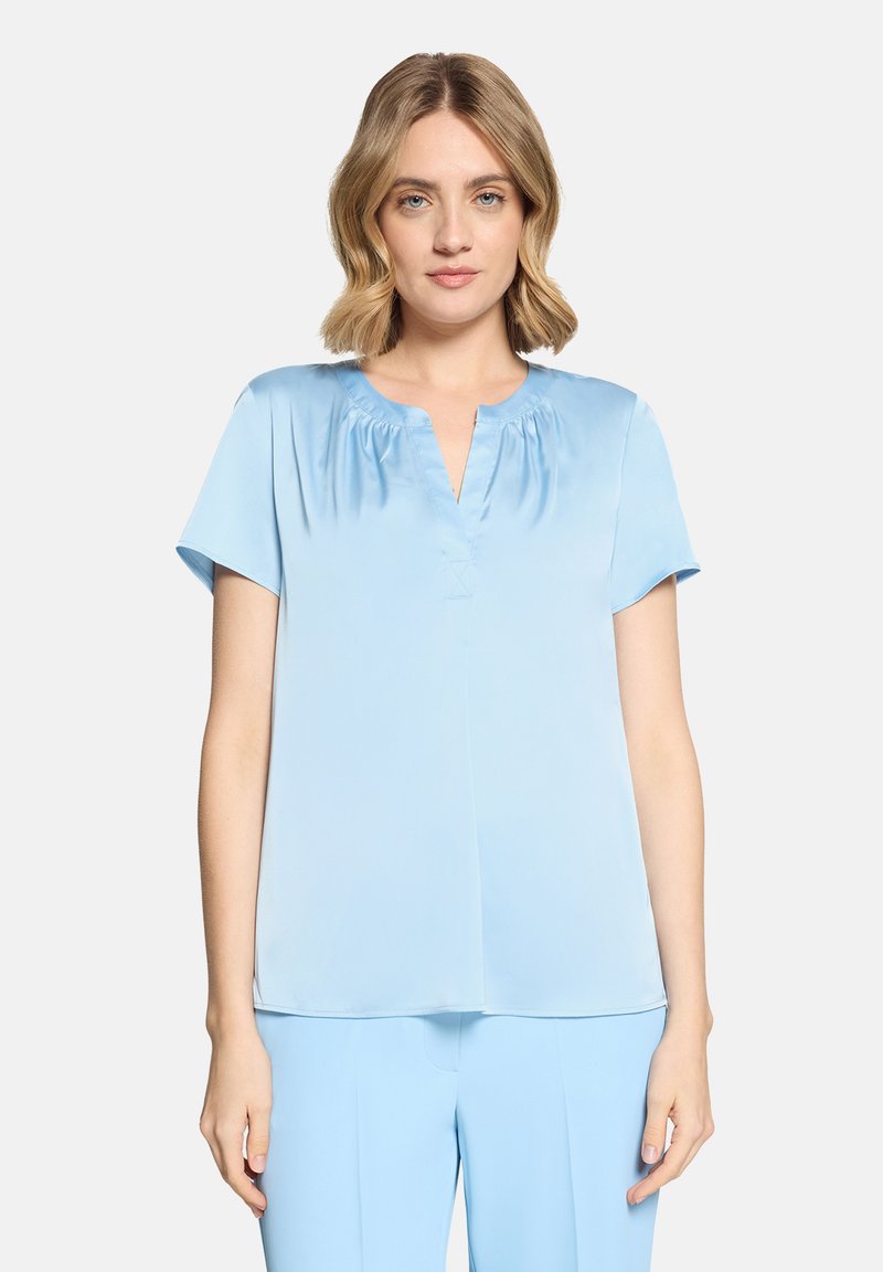 Femme aux cheveux blonds mi-longs portant un chemisier bleu clair à manches courtes et un pantalon assorti bleu clair, debout devant un fond uni.