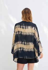 Vêtement de style kimono tie-dye noir et beige, ample avec de larges manches et un ourlet arrondi. Texture douce du tissu et absence de fermetures.