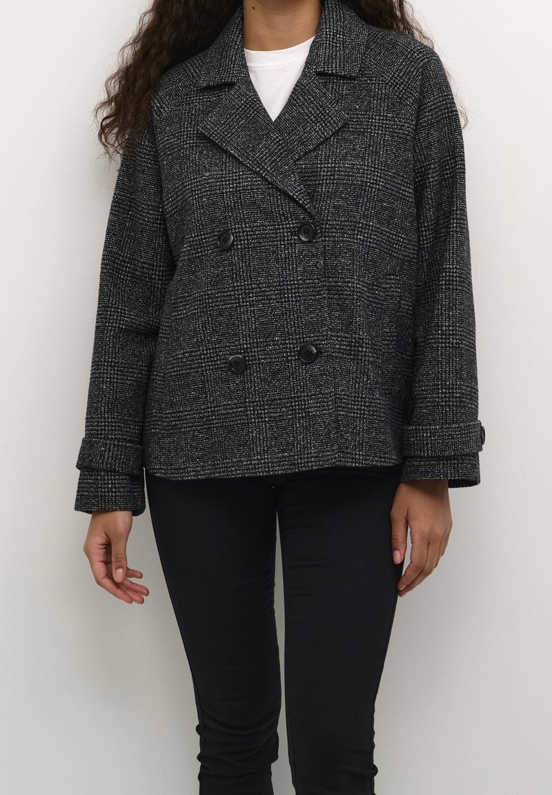 Personne portant une veste croisée à carreaux noirs et gris avec des boutons noirs et un pantalon noir sur un fond uni.