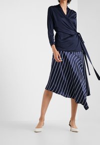 Námořnický zavinovací top z hladkého materiálu s dlouhými rukávy, spojený s asymetrickou sukní po kolena s bílými vertikálními pruhy. Bílé podpatky.