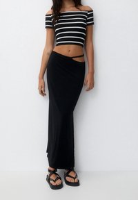 Černý crop top s odhalenými rameny a pruhy, černá maxi sukně s bočním výřezem a černé sandály s tenkými pásky. Hladké, přiléhavé materiály.