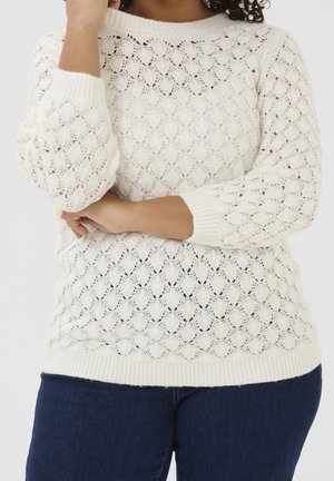 Personne portant un pull blanc en tricot avec un motif en festons et un jean bleu foncé, les bras croisés à la taille et touchant le menton.