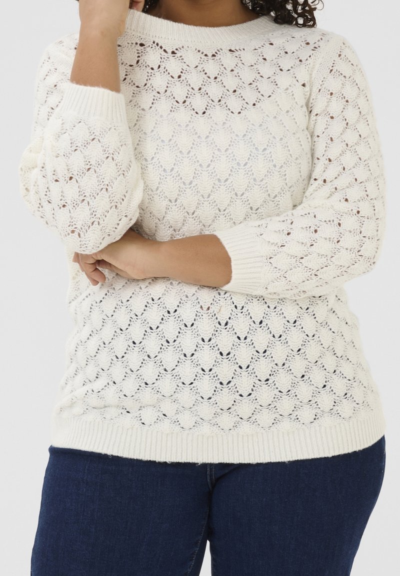 Personne portant un pull blanc en tricot avec un motif en festons et un jean bleu foncé, les bras croisés à la taille et touchant le menton.