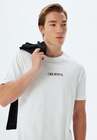 Hvid bomulds t-shirt med ribbet halsudskæring og korte ærmer, med sort broderet tekst "GREATFUL" på brystet. Sort beklædning hængende over skulderen.