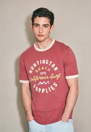 Ung mand med mørkt hår iført en rød t-shirt med hvide kanter og teksten California surf, hænderne i lommerne på lyseblå jeans, neutral baggrund.