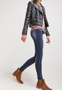Veste de motard en cuir noir avec des garnitures en métal argenté, haut à motifs, jeans skinny en denim bleu et bottines marron à la finition lisse.