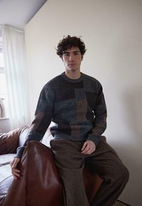 Pullover mit einem Patchwork-Design in dunkelgrauen, blauen und braunen Farbtönen, aus strukturiertem Stoff. Braune Schlaghosen und ein Lederstuhl.