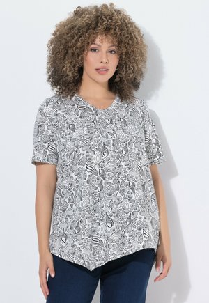 Frau mit lockigem Haar, die eine kurzärmelige weiße Bluse mit schwarzen Muschelmuster und dunkelblaue Hosen vor einem einfarbigen hellen Hintergrund trägt.