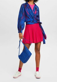 Blouse cache-cœur bleue avec motif abstrait coloré, associée à une jupe plissée rose, un sac tissé bleu et des baskets blanches avec des accents roses.