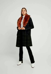 Zwarte geruite jas met een grote rood-bruin nepbont kraag, gecombineerd met een witte coltrui, zwarte broek en witte haklaarzen.