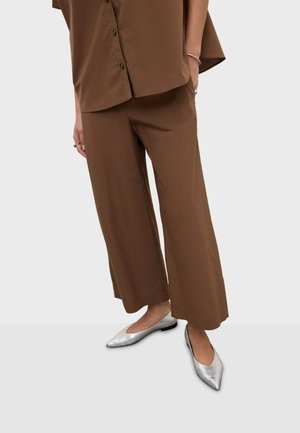 Osoba ubrana w brązowe, szerokie spodnie 7/8, brązową koszulę na guziki oraz srebrne, spiczaste baleriny, stojąca na jednolitym tle.