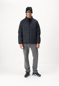 Granatowa kurtka puchowa z poziomym pikowaniem, szare dżinsy z przednimi kieszeniami i czarne buty sportowe. Dodatki to czapka beanie w kolorze czarnym oraz okulary przeciwsłoneczne.