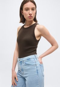 Γυναίκα με κοντά καστανά μαλλιά που φοράει αμάνικο σκούρο καφέ τοπ και ανοιχτόχρωμα μπλε τζιν, ποζάροντας με το ένα χέρι στην τσέπη της.