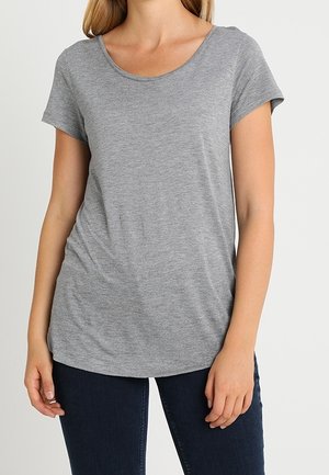 Frau trägt ein schlichtes graues T-Shirt mit kurzem Ärmel und Rundhalsausschnitt sowie dunkelblaue Jeans, steht mit entspannten Armen an der Seite.