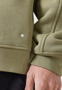 Sudadera de color verde oliva con puños acanalados, que presenta un pequeño logotipo plateado en la muñeca izquierda. Textura suave y ajuste relajado visibles.