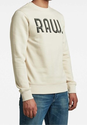 Homme portant un sweat-shirt crème à manches longues avec "RAW." en lettres larges et foncées, assorti à un jean en denim bleu.