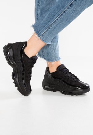 Osoba nosząca czarne sportowe buty z grubymi podeszwami i siateczkowymi detalami, w połączeniu z niebieskimi, pranymi, skróconymi dżinsami, stojąca na białej podłodze.