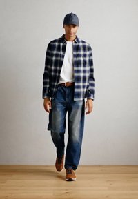 Chemise à carreaux bleu marine et crème, t-shirt blanc, jeans bleus et baskets marron ; modèle portant une casquette grise. Fond en bois.