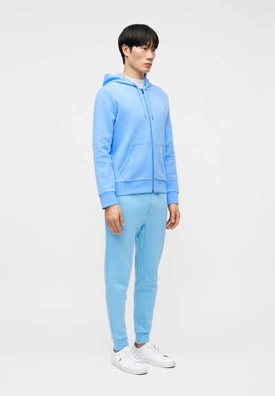 Νεαρός άνδρας όρθιος, φορούσε ανοιχτό μπλε φούτερ με φερμουάρ και αντιστοιχισμένο παντελόνι φόρμας, λευκά αθλητικά παπούτσια, κοιτάζοντας προς τα δεξιά σε απλό γκρι φόντο.