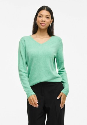 Groene v-hals trui van zacht materiaal, met lange mouwen en ribbenmanchetten. Gecombineerd met zwarte high-waisted broek, minimalistisch ontwerp.
