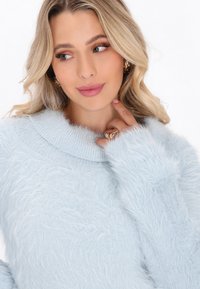 Maglione azzurro chiaro e morbido, con un ampio colletto ripiegato e maniche lunghe. Texture soffice e vestibilità rilassata. Accentuato da un anello su una mano.
