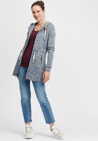Abbigliamento esterno con cappuccio in tessuto a maglia blu con lacci bianchi, dotato di due tasche frontali, abbinato a una maglia bordeaux e jeans.