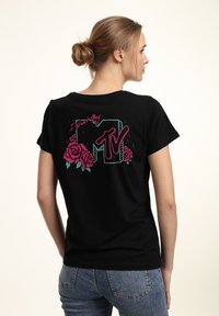 Sort bomulds T-shirt med et farverigt grafisk tryk bagpå, der viser roser og teksten "I Want My MTV" i pink og grøn.